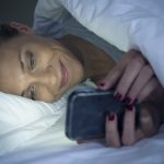 Kvinde ligger i sin seng og bruger netdating på sin iPhone.